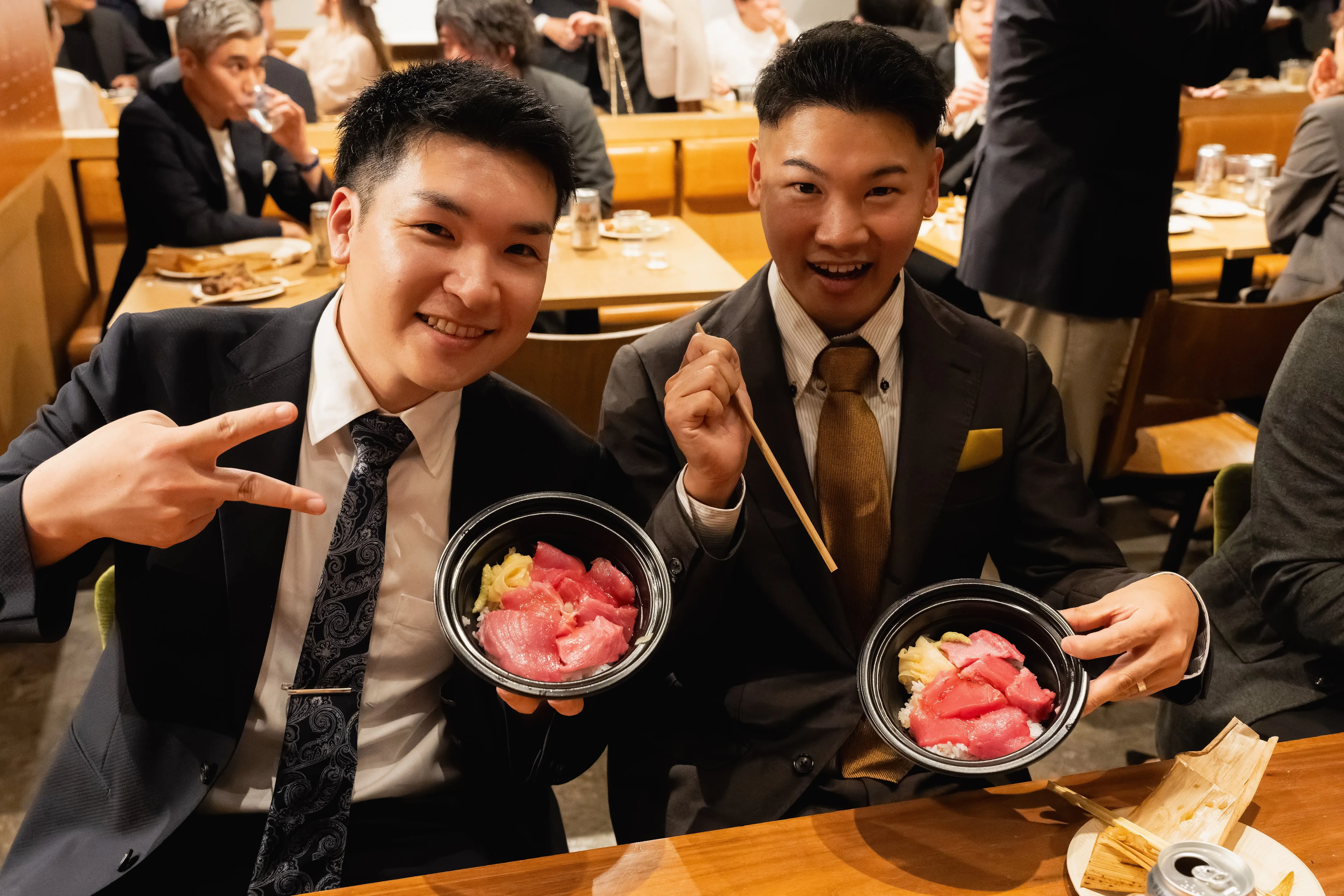 スーツ姿の参加者2名が海鮮丼を手に笑顔を見せる場面